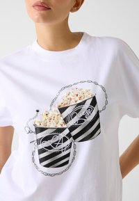 Wit T-shirt met een afbeelding van twee popcornbakjes, zwart-wit gestreept, gevuld met popcorn en verbonden door een ketting. Zilverkleurige accenten zichtbaar.