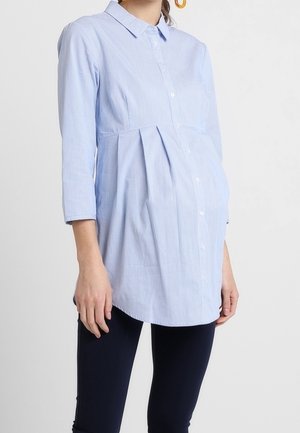 Femme enceinte portant une chemise de maternité bleu clair à boutons et un pantalon foncé, debout sur un fond blanc uni.
