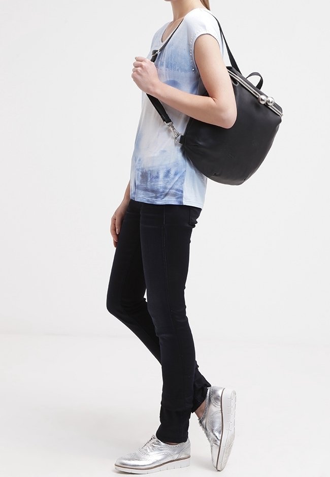 Personne portant un pantalon noir et une chemise tie-dye bleue, portant un sac à bandoulière en cuir noir avec un fermoir argenté, debout sur fond blanc.
