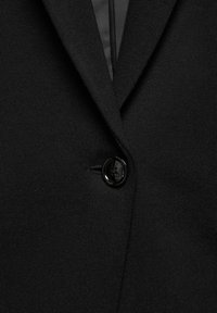 Blazer noir à la texture lisse, doté d'un col à cran classique et d'une fermeture à un seul bouton noir. Design simple et ajusté.