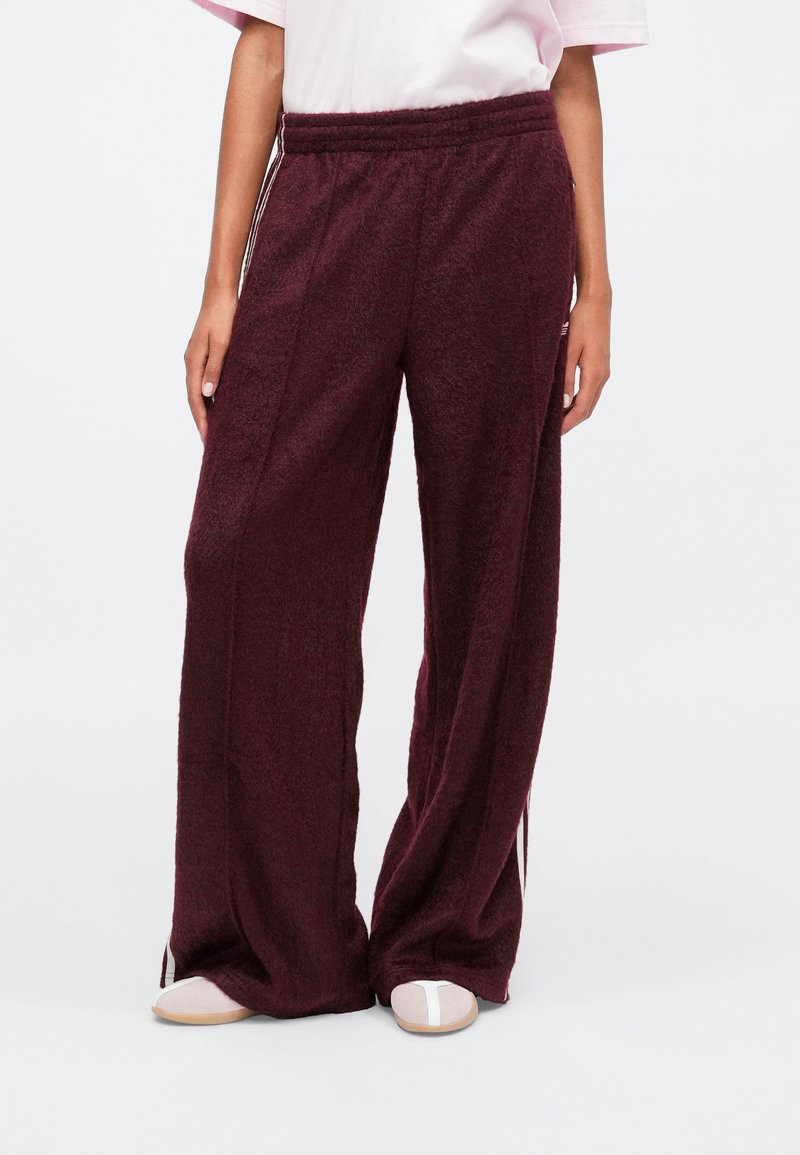 Pantaloni a gamba larga color bordeaux in morbido tessuto testurizzato, con vita elastica e strisce bianche laterali. Abbinati a scarpe chiare.