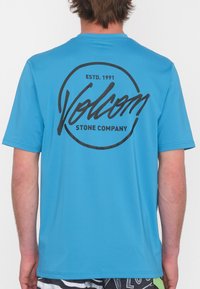 Camiseta de manga corta azul con un logo circular negro y texto en la parte de atrás. Textura suave, diseño de cuello redondo.
