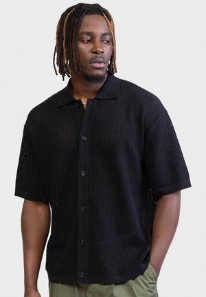 Άνδρας με μαύρες και καφέ πλεξούδες, φοράει μαύρο κοντομάνικο πουλόβερ με κουμπιά και ελαιί πράσινο παντελόνι, κοιτάει προς τα πλάγια.