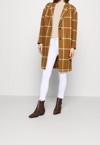 Abrigo de lana a cuadros marrón con mangas largas, top beige, jeans skinny blancos y botines marrones con acentos texturizados.