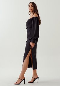 Schwarzes Off-Shoulder-Kleid mit langen, puffigen Ärmeln und einem Schlitz bis zum Oberschenkel. Schmaler Schnitt, aus weichem Stoff gefertigt. Kombiniert mit schwarzen Riemchen-Heels.