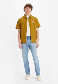 Short-sleeve mustard button-up shirt with a leaf embroidery, paired with light blue jeans and beige sneakers, featuring a relaxed fit.