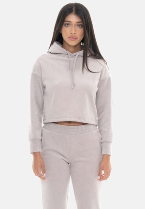 Junge Frau steht und trägt einen hellgrauen kurz geschnittenen Hoodie mit Kordeln und passende hoch taillierte Hosen vor einem schlichten Hintergrund.