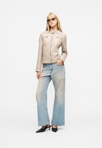 Ξανθιά γυναίκα με μπεζ ζακέτα με φερμουάρ, ανοιχτό μπλε τζιν με φαρδιά μπατζάκια, μαύρα τακούνια και γυαλιά ηλίου, στέκεται με το ένα χέρι στην τσέπη.