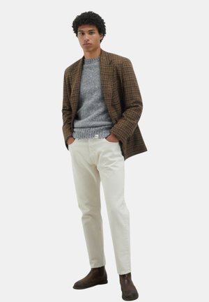Un giovane uomo sta in piedi con le mani nelle tasche, indossando un blazer a quadri marrone, un maglione grigio, pantaloni crema e stivali marrone scuro su sfondo bianco.