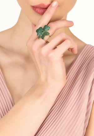 Gold-tone ring featuring a green glitter leaf design, placed on a finger, with a soft pink pleated fabric in the background.