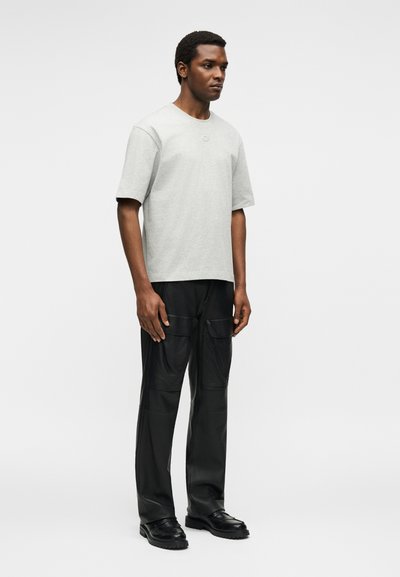 Άνδρας στέκεται φορώντας ένα ανοιχτό γκρι κοντομάνικο μπλουζάκι, μαύρο παντελόνι cargo με τσέπες και μαύρα παπούτσια σε απλό ανοιχτό φόντο.