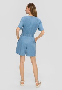 Combinaison en denim à manches courtes, taille élastique et poches. Couleur bleu clair avec une coupe décontractée et des shorts retroussés. Vue arrière montrée.