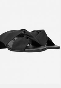 Zwarte satijnen slidesandalen hebben gekruiste banden met een geknoopte uitstraling, open neus en een platte rubberen zool. De textuur lijkt glad en zacht te zijn.