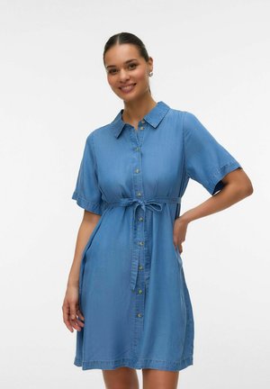 Kobieta uśmiechająca się i ubrana w niebieską, dżinsową sukienkę na krótki rękaw z guzikami i paskiem zawiązywanym w talii, na jednolitym białym tle.
