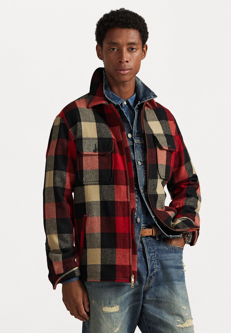 Red, black, and beige plaid jacket with a zip front, two chest pockets, and adjustable collar, layered over a denim shirt and distressed jeans.