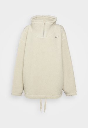 Kremowy polarowy pulower z półzamkiem, wysokim kołnierzem, długimi rękawami, regulowanymi troczkami u dołu oraz małym czarnym logo Nike na piersi.
