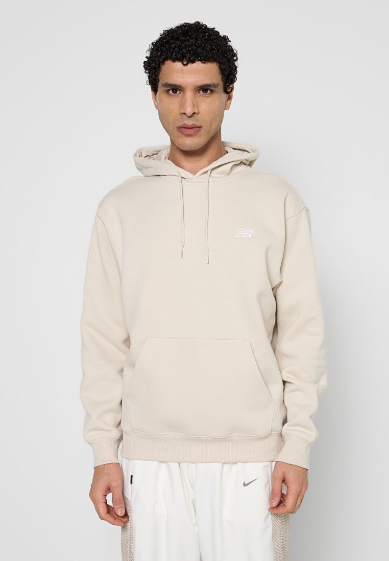 Mann mit lockigem Haar, der einen beigen New Balance Hoodie und weiße Nike-Hosen trägt und vor einem schlichten hellgrauen Hintergrund steht.