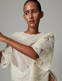 Κρεμ διάφανο τοπ με κυματιστή υφή υφάσματος, κοντά μανίκια και λεπτομέρειες με ανοίγματα. Ο σχεδιασμός διαθέτει στρωματοποιημένα κρόσια για διάσταση.