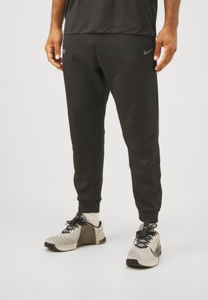 Pantalones deportivos atléticos negros con piernas cónicas, puños elásticos, bolsillos laterales y un pequeño logo metálico de Nike en la parte superior del muslo izquierdo.