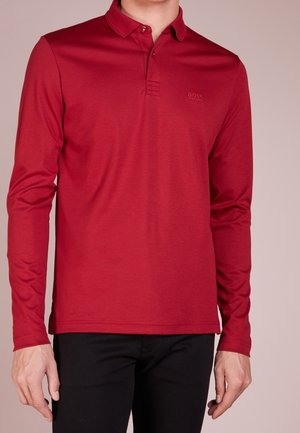 Mand iført en rød poloshirt med lange ærmer, krave og knapper, kombineret med sorte bukser mod en neutral baggrund.