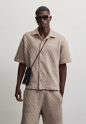 Homme portant une chemise beige texturée à manches courtes et un short assorti, un sac bandoulière noir, des lunettes de soleil foncées et un collier en or sur un fond uni.