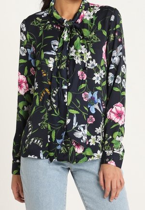 Woman wearing a navy floral blouse with a front tie neck and long sleeves, paired with light blue jeans.