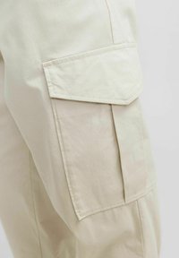 Pantalones cargo beige con tejido texturizado, bolsillos laterales con solapas y detalles de costura visibles. Diseñados para utilidad y comodidad.