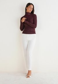 Burgundské rolákové triko s texturovaným povrchem, spárované s bílými těsnými kalhotami. Modelka má na sobě bílé sandály na otevřenou špičku. Neutrální pozadí.