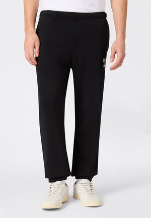 Uomo che indossa pantaloni sportivi con vita elastica neri con logo bianco sulla coscia, abbinati a scarpe da ginnastica bianche e calzini bianchi su sfondo neutro.