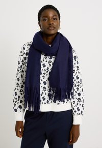 Bufanda de color marino hecha de tela suave con flecos largos, llevada sobre un sudadero blanco con un estampado de leopardo en negro y azul.