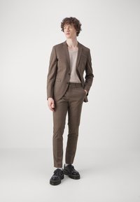 Costum cu carouri maronii, cu sacou slim fit și pantaloni asortați. Cămașă tricotată crem dedesubt, asociată cu pantofi negri cu talpă masivă.
