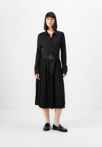 Svart skjortekjole med lange ermer og plissert skjørt, med et tettsittende belte i midjen og knappelukking. Båret med svarte loafers og beige sokker.