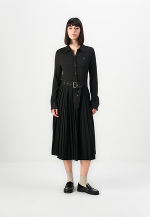 Svart skjortekjole med lange ermer og plissert skjørt, med et tettsittende belte i midjen og knappelukking. Båret med svarte loafers og beige sokker.