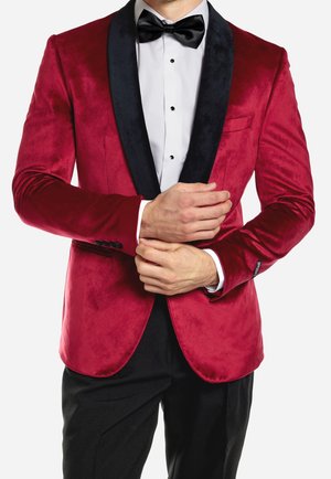 Man die zijn mouwen aanpast en een rood fluwelen smokingjasje met zwarte revers, een wit overhemd, een zwarte strik en zwarte broek draagt.