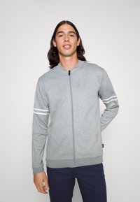 Chaqueta gris con cierre zip y cuello alto, con rayas blancas en las mangas y puños acanalados. Material suave, diseño minimalista.