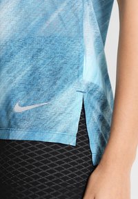 Modrý sportovní top s výrazným vzorem, vyrobený z prodyšné látky, s bočními rozparky a malým stříbrným logem Nike u lemu. Doplněno strukturovanými černými legínami.