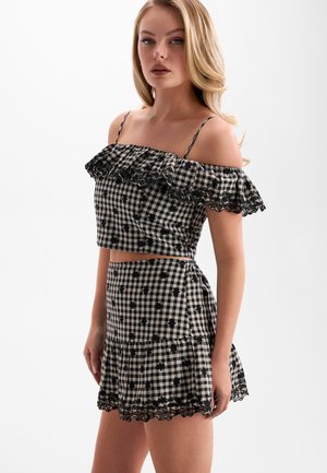 Vrouw draagt een zwart-wit geruit off-shoulder cropped top en bijpassende rok met kanten rand en kleine bloemenpatronen.