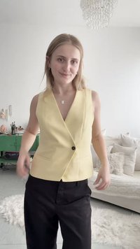 Νέα γυναίκα με ξανθά μαλλιά, που φοράει αμάνικο κίτρινο γιλέκο και μαύρο παντελόνι, στέκεται σε ένα μοντέρνο σαλόνι με ένα λευκό καναπέ.