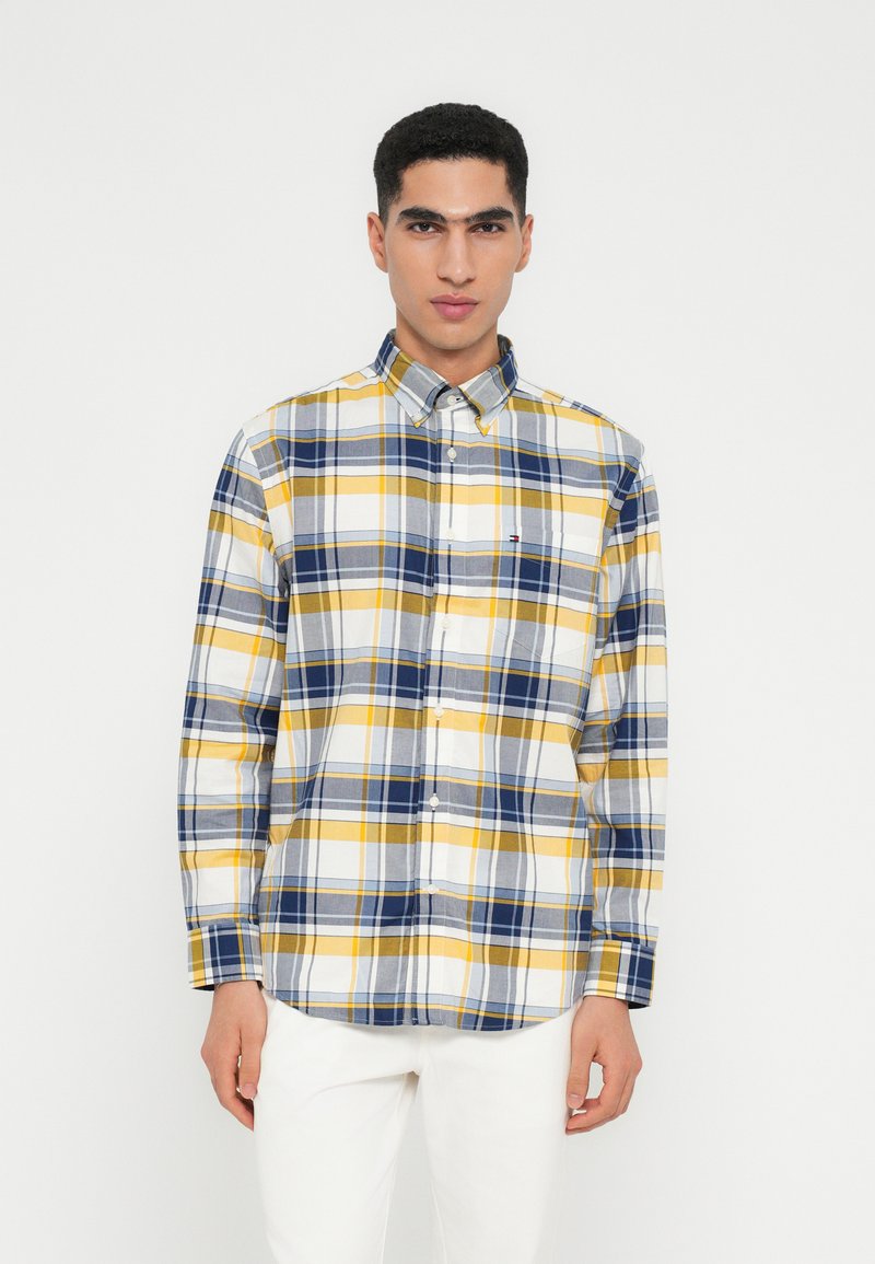 Mann in einem Langarmhemd mit blauem, gelbem und weißem Karomuster und weißen Hosen, der vor einem einfarbigen Hintergrund steht.