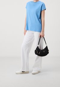 Svart nylonaxelväska med guldfärgad hårdvara, triangulär logotyp och dragkedjor. Bärs med en ljusblå topp och vita vida byxor. Vita skor.