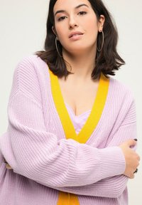 Cardigan a maglia lavanda con scollo a V giallo, maniche lunghe e polsini a coste. Indossato sopra un top viola chiaro. Tessuto aderente e texturizzato.