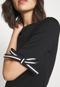 Černý top s krátkým rukávem, který se vyznačuje texturovanou tkaninou a jedinečným designem rukávů, s černobílými pruhovanými stužkami uvázanými na lokti.