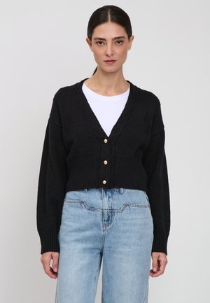 Donna con capelli scuri che indossa un cardigan nero con bottoni sopra una canottiera bianca e jeans azzurri a vita alta, in piedi davanti a uno sfondo semplice.