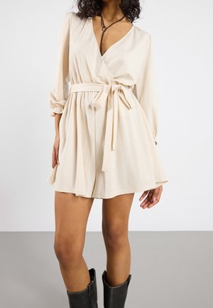 Vrouw draagt een crèmekleurige wikkeljurk met een gestrikte ceintuur, gecombineerd met zwarte laarzen tot halverwege de kuit, staand tegen een effen achtergrond.