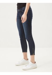 Jeans de mezclilla azul oscuro con corte ajustado, de longitud hasta el tobillo, con un detalle de cremallera en el dobladillo y bolsillos estándar. Llevados con zapatillas blancas.