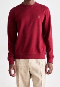 Sweat-shirt rouge à manches longues, poignets et ourlet côtelés, avec un petit patch logo. Confectionné en coton lisse. Se marie avec un pantalon de couleur claire.