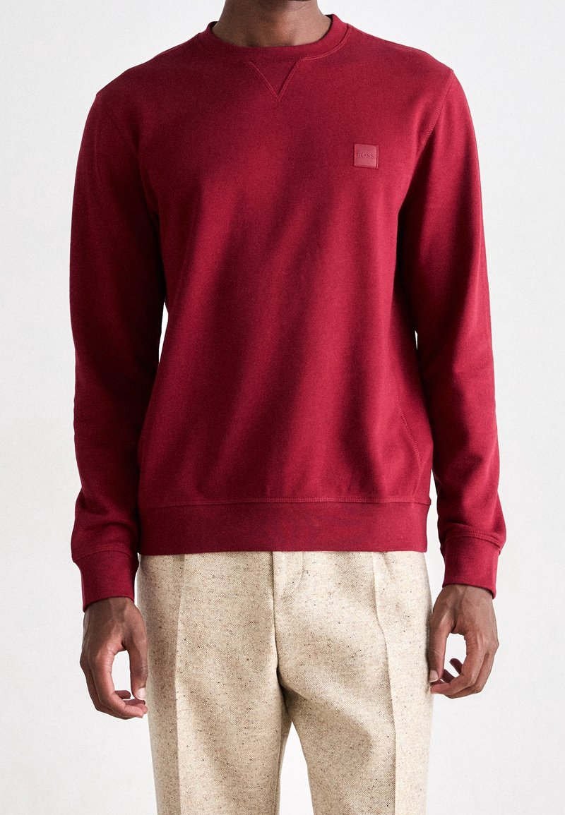 Sweat-shirt rouge à manches longues, poignets et ourlet côtelés, avec un petit patch logo. Confectionné en coton lisse. Se marie avec un pantalon de couleur claire.