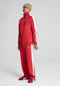 Rød atletisk træningsdragt med hvide side-striber. Jakke med lynlås, matchende bukser og røde sko. Fremstillet af glat, strækbart stof.