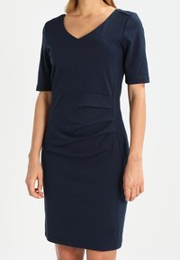 Mörkblå, figurnära klänning med korta ärmar, v-ringning, texturerade sidopaneler och rynkade detaljer i midjan för en skräddarsydd look.