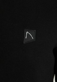 Maglione nero a maglia con tessuto testurizzato, dotato di un'etichetta quadrata con un emblema grafico bianco. Design semplice con scollatura rotonda.